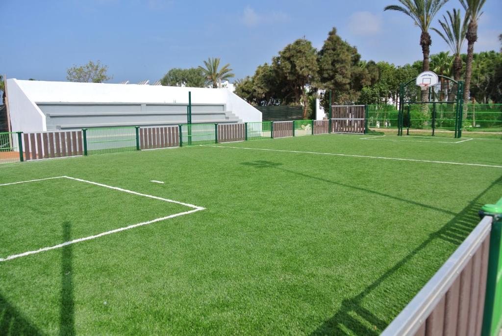 Mākslīgā zāle sporta laukumā ar futbola laukuma marķējumu un basketbola grozu, apkārt žogs un zaļi koki fona.