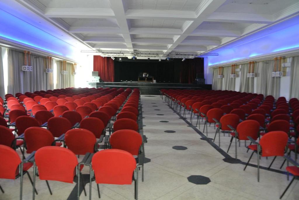 Sarkano krēslu rinda konferences zālē ar skatuvi, gatava pasākumam. Zāles apgaismojums rada patīkamu atmosfēru.