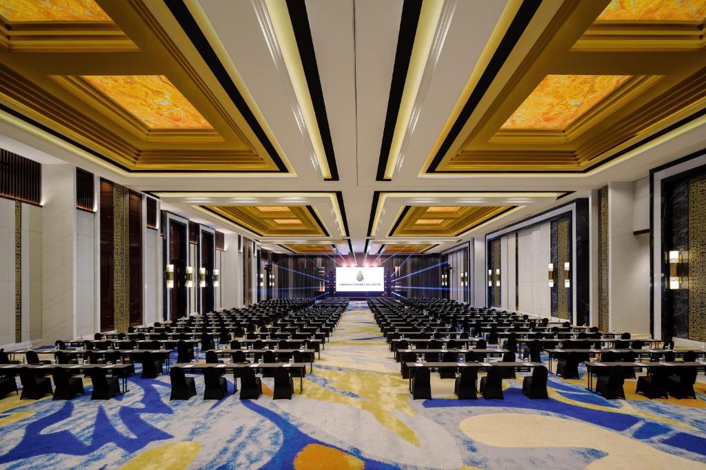 Liela, moderna konferences telpa ar elegantiem griestiem un rindiņām melnu galdu, kas gatavi pasākumam. Centrā atrodas ekrāns prezentācijām, radot profesionālu atmosfēru.