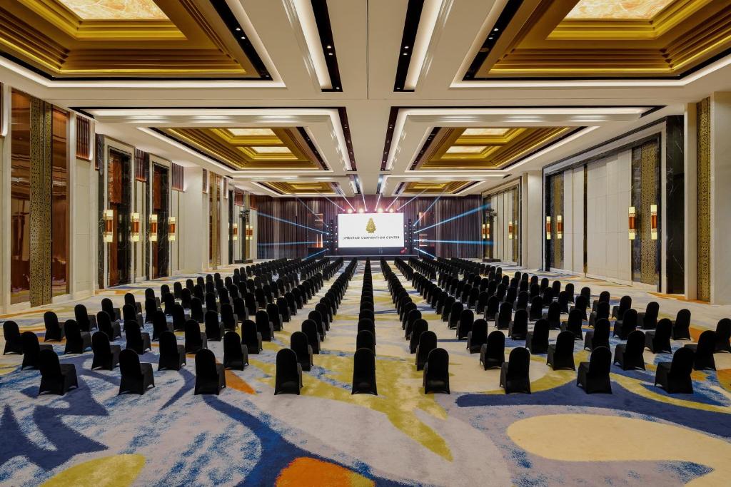 Mūsdienīga konferences zāle ar kārtīgi novietotām melnām krēslām un lielu ekrānu priekšā, kas paredzēta sanāksmēm un pasākumiem. Telpa ir elegantā dizainā ar greznām griestiem un krāsainu paklāju.