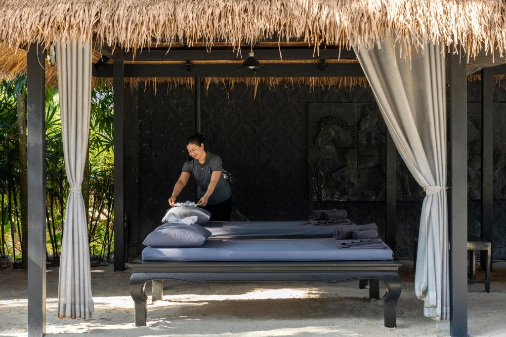 Spa procedūru telpa ar diviem masāžas galdiem, kur darbiniece kārto palagus. Apkārt ir zaļa vide un dekoratīvas detaļas, kas radījušas relaksējošu atmosfēru.