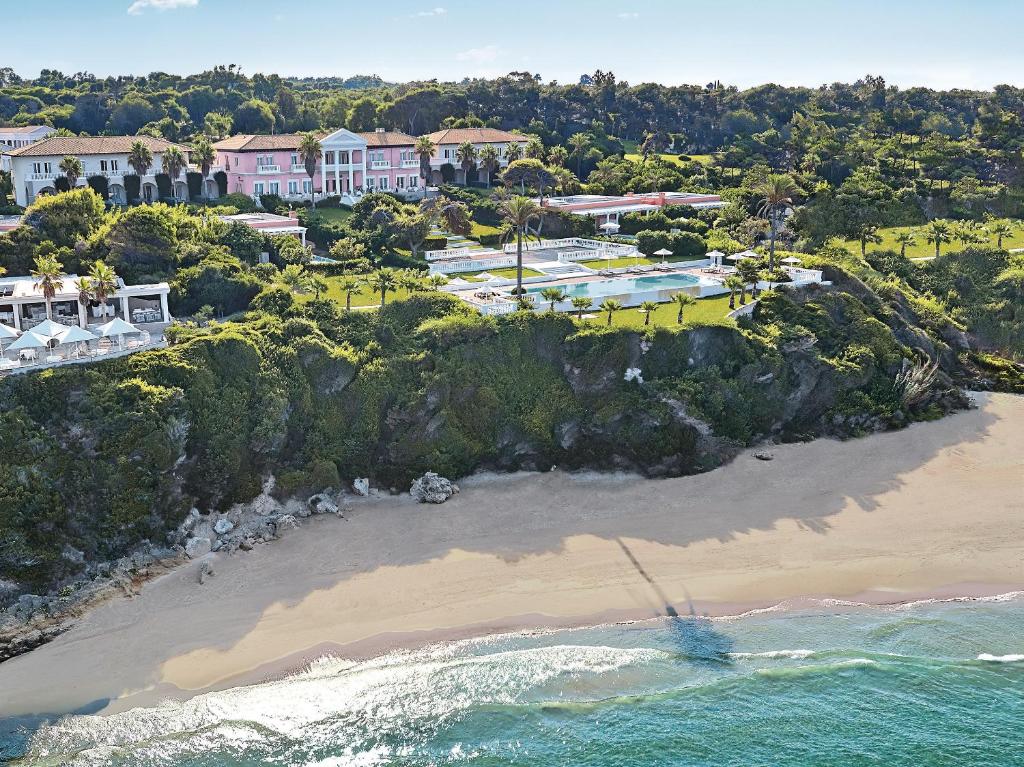 Vistas uz greznu villu, kas atrodas kalnā pie pludmales, ar baseinu un zaļu ainavu. Ūdens krastā ir smiltis, un attēls piedāvā relaksējošu atmosfēru.