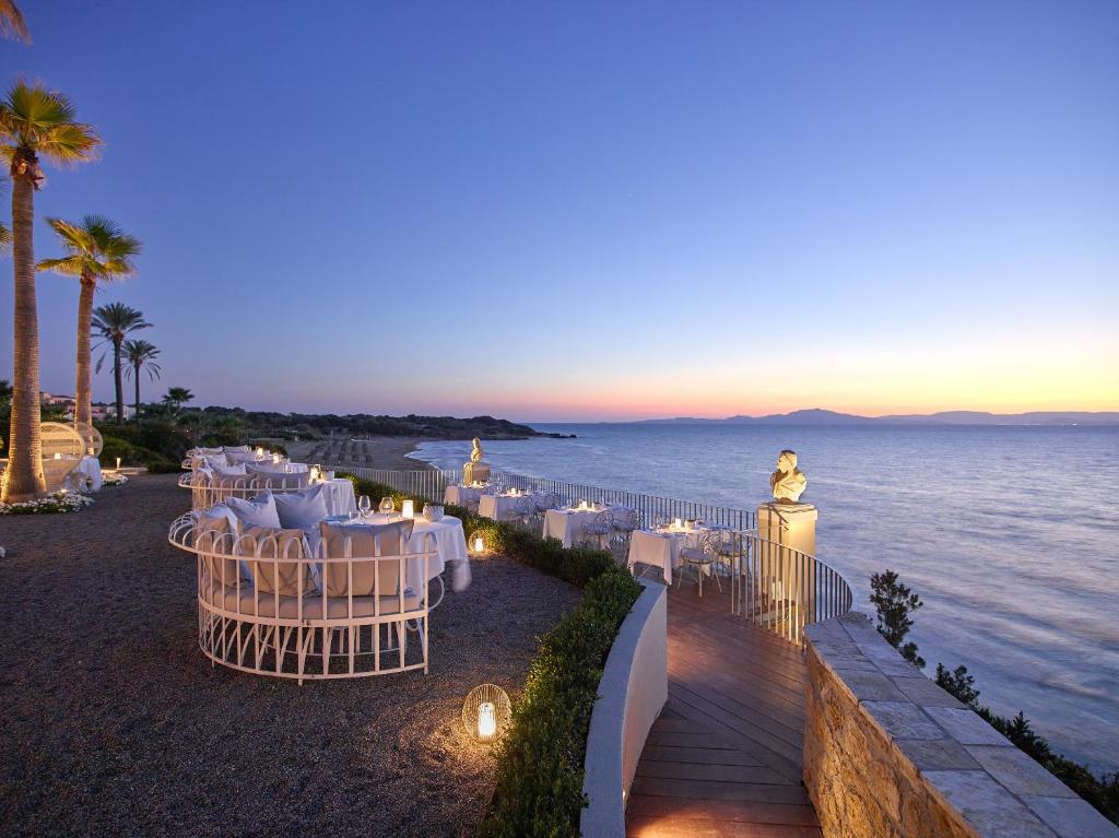 Elegantā pludmales restorāna terase, apgaismotā ar mīkstu gaismu, piedāvā romantisku skatu uz jūru saulrieta laikā. Baltas mēbeles un palmas rada relaksējošu atmosfēru.