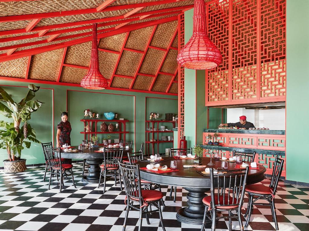 Elegants restorāns ar melnbaltu grīdu un krāsainu interjeru, kurā redzami tradicionāli elementi un gaismas ķermeņi. Galdi ir klāti ar galda piederumiem, bet fons piepildīts ar dekoratīvām detaļām un augiem.