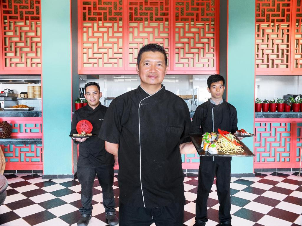 Trīs pavāri stāv restorānā, turot plāksnes ar garšīgām ēdienu porcijām. Interjers ir krāsains, ar rakstainām sienām un flīzēm.