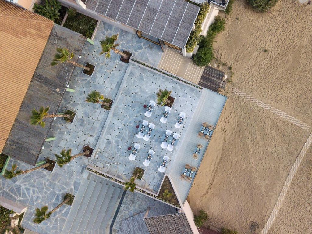 Pārskats no gaisa uz modernu terasi ar koka un akmens elementiem, kur izvietoti galdi un krēsli, apkārt aug palmām. Blakus redzama smilšu pludmale.