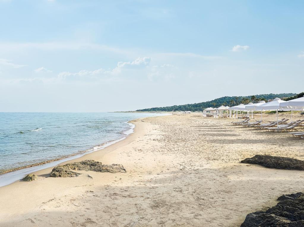 Pludmale ar smiltīm un mierīgu jūru, kurā redzami sauļošanās krēsli un baldahīni. Šī ainava rada relaksējošu un mierīgu atmosfēru.