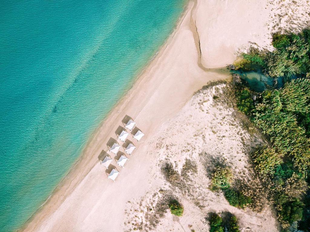 Pludmales ainava ar baltām saulessargiem, kas izvietoti pie kristāldzidra ūdens un smilšu krasta, ko ieskauj zaļumi.