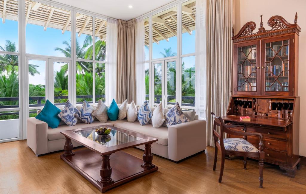 Mūsdienīgs viesistabas interjers ar plašiem logiem, kas paver skatu uz zaļām palmām. Stūra dīvāns ar spilveniem un elegants koka rakstāmgalds pie loga rada mājīgu atmosfēru.