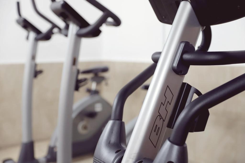 Trenažieru zāles skats ar diviem moderniem trenažieriem, ieskaitot eliptisko trenažieri un velosipēdu, uz neitrālas krāsas fona.