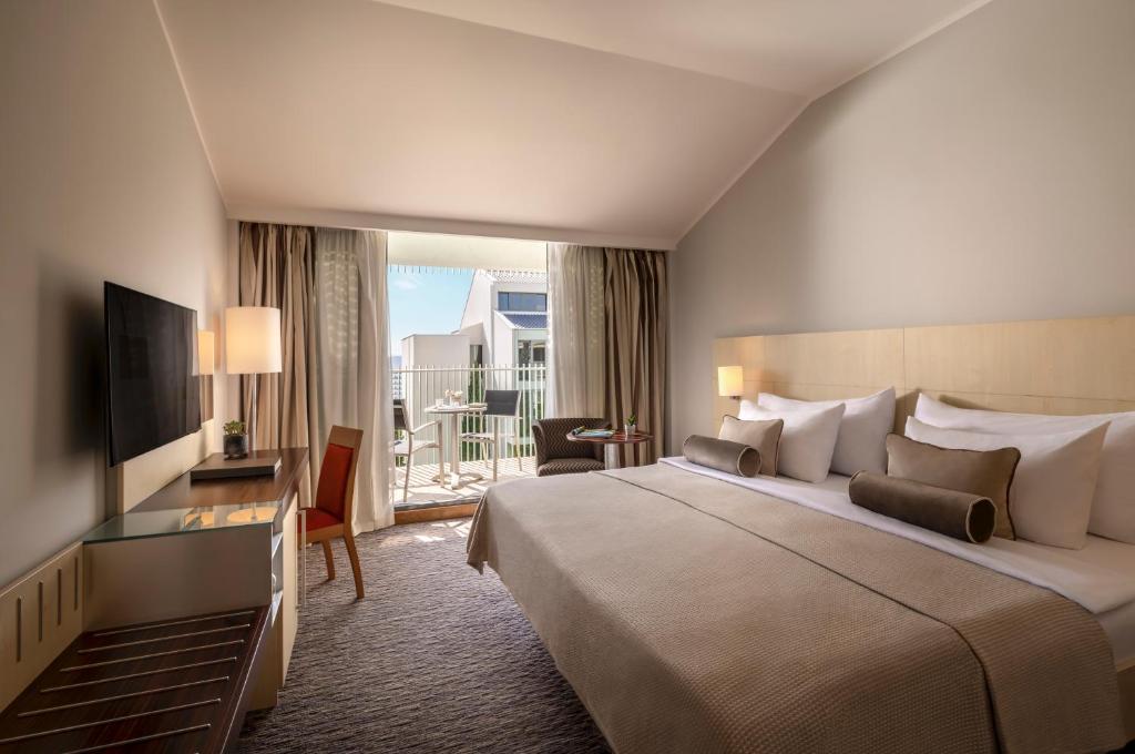 Mūsdienīga viesnīcas numura iekārtojums ar plašu gultu, televizoru un balkonu, no kura paveras skats uz ārpusi. Interjers ir gaišs un elegants, radot komfortablu atmosfēru.