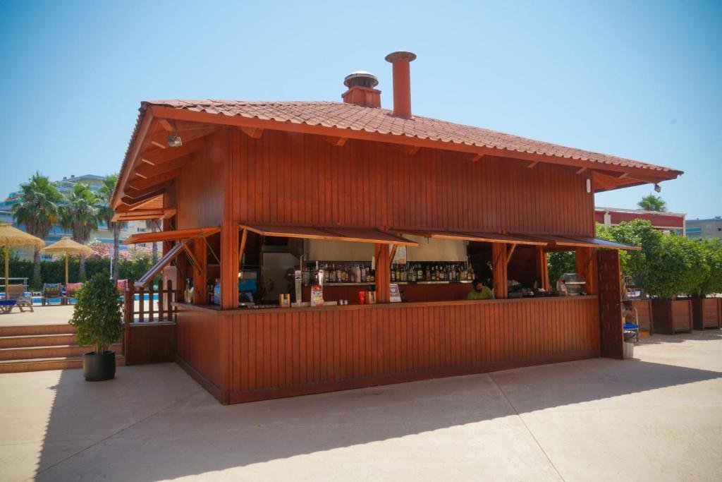Koka bārs ar sarkanu jumtu, kas atrodas pie peldbaseina, apkārt palmām un sauļošanās krēsliem. Bāra priekšpusē redzamas daudzas dzērienu un ēdienu izvēles.