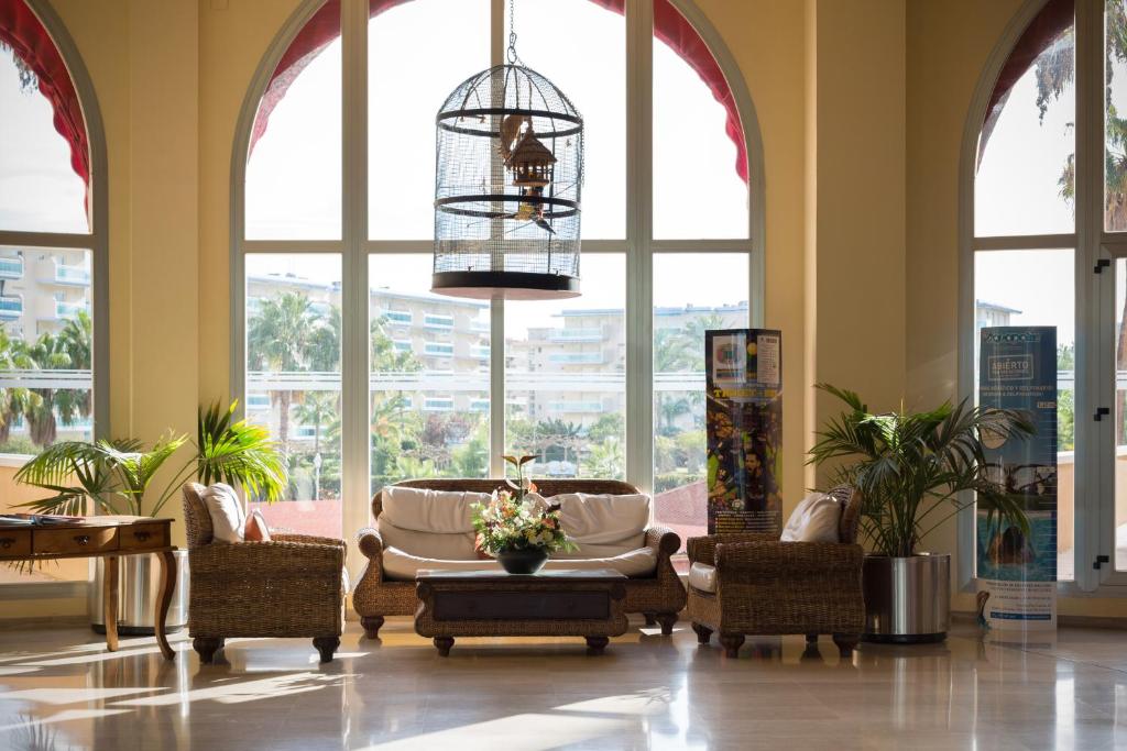 Gaiša un plaša viesnīcas halle ar ērtām mēbelēm, dekoratīvām augu kompozīcijām un gaisīgu putnu būri, kas karājas pie loga. Skats uz zaļo apkārtni caur lielajiem logiem.