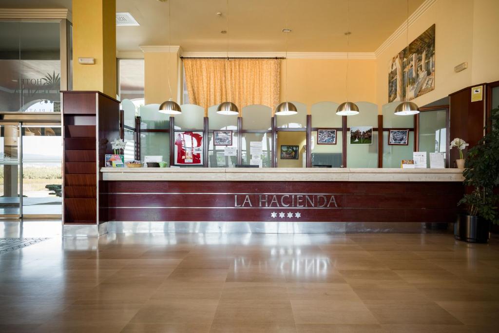 Reģistratūra viesnīcā "La Hacienda" ar modernu dizainu, koka un stikla elementiem, apgaismota ar stilīgām lampām. Uz galda redzami dažādi informācijas materiāli un augu kompozīcija.