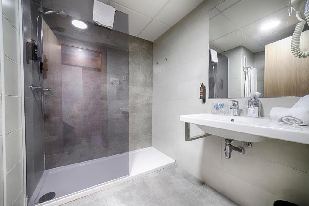 Mūsdienīga vannas istaba ar stiklotu dušu, izsmalcinātu izskatu un gaišām flīzēm, kas nodrošina komfortu un stilīgu dizainu. Dušas stūrī ir redzamas dvieļi un higiēnas piederumi.