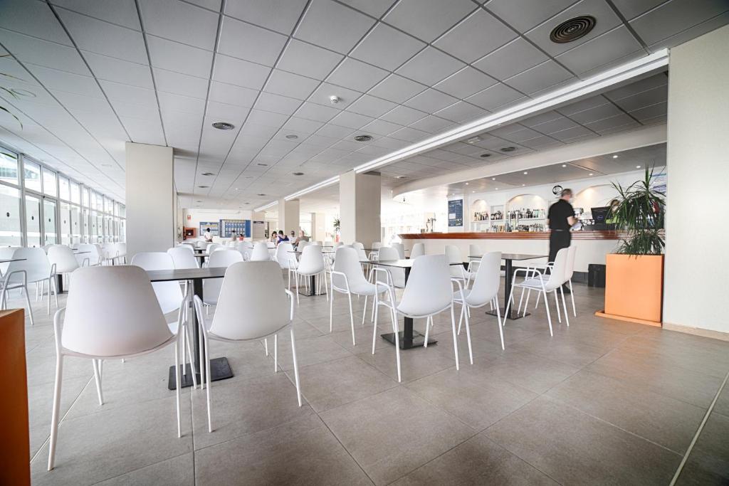 Mūsdienīgs kafejnīcas interjers ar baltām krēslām un galds, gaišs apgaismojums un plašas logu ailes, kas nodrošina dabīgu gaismu. Klientu un darbinieku klātbūtne rada dzīvīgu atmosfēru.