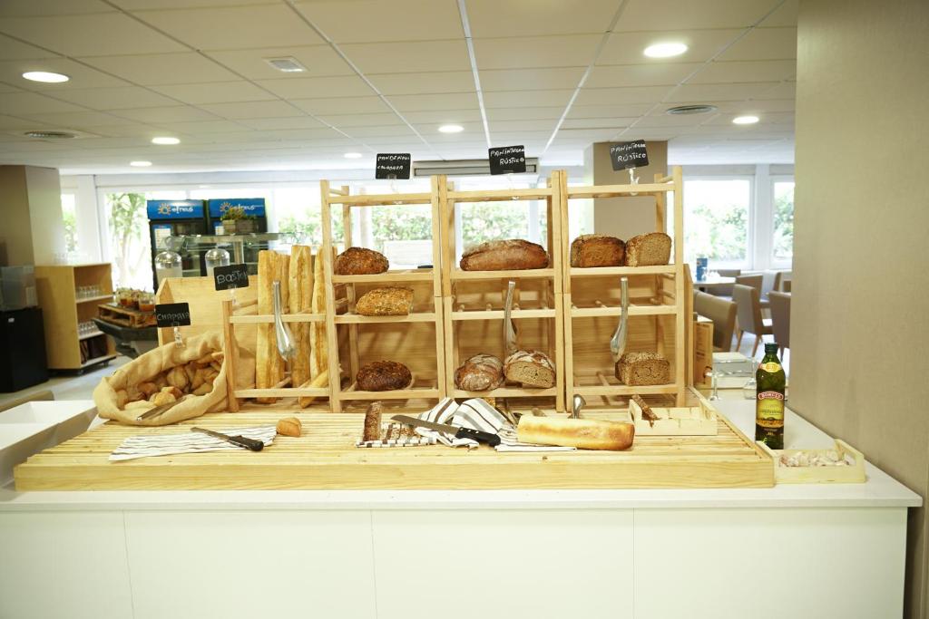 Maizes stends ar dažādām maizes šķirnēm un cepumiem, kas sakārtoti koka plauktā. Fonā redzama gaisīga telpa ar ēdināšanas zonu.