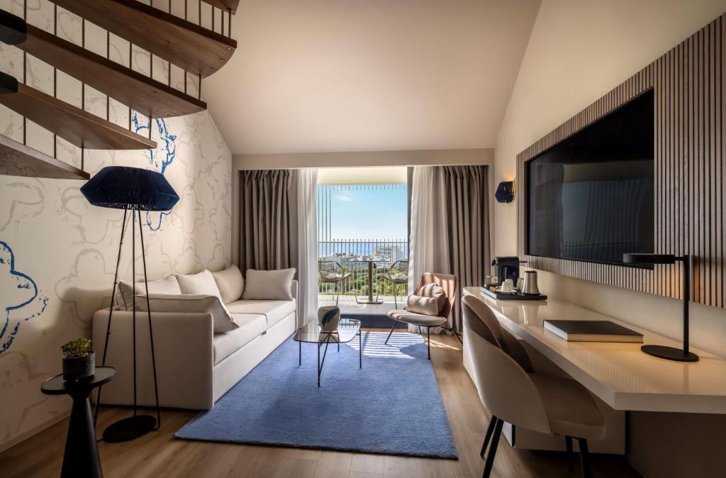 Mūsdienīgs viesnīcas numurs ar balkonu un skatu uz ainavu, iekārtots ar ērtu dīvānu, rakstāmgaldu un stilīgu apgaismojumu. Interjers ir gaišs un elegants, radot relaksējošu atmosfēru.