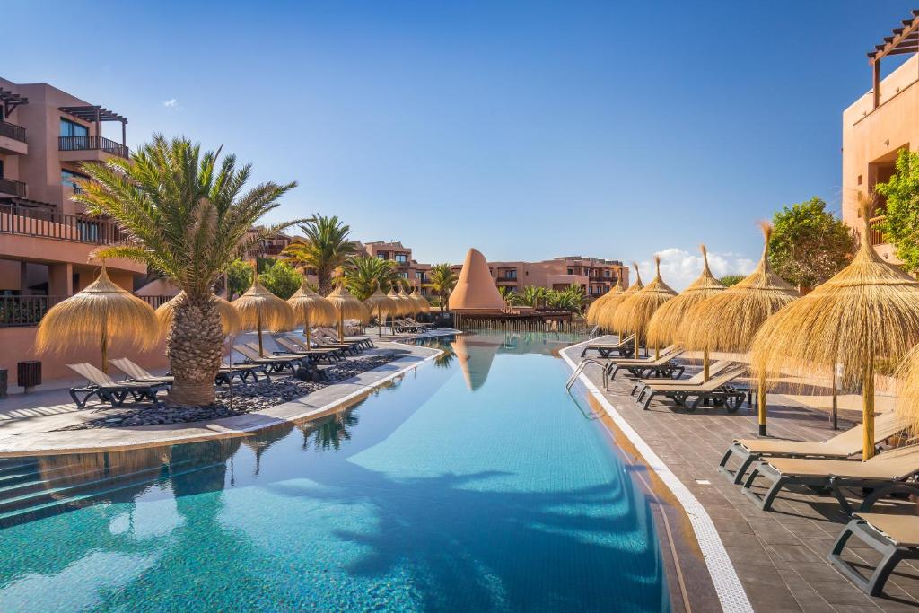 Luksusa viesnīcas peldbaseins ar palmām un sauļošanās krēsliem, ieviests tropiskās atmosfēras elements. Apkārtne ir labi kopta, ar skatu uz zilo debesu fonu.