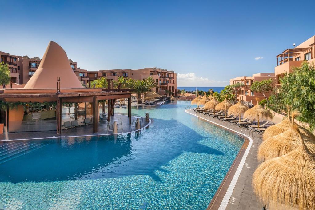 Luksusa viesnīcas peldbaseins ar modernu arhitektūru un palmām, kas rada tropisku atmosfēru. Skats uz jūru un saulainu debesi papildina relaksējošo vidi.