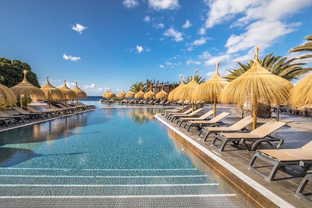 Luksusa viesnīcas baseins ar sauļošanās krēsliem un salmiem pārklātiem nojumes. Skaista debesis un zils jūras skats papildina atpūtu.