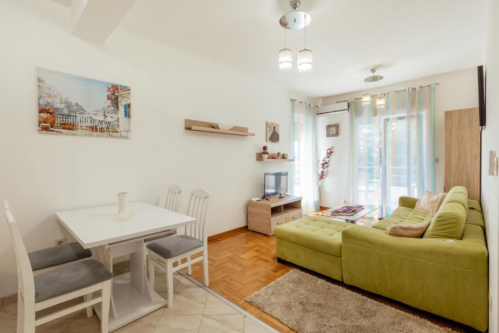 Mūsdienīgs viesistabas iekārtojums ar zaļo dīvānu, ēdamgaldu un televizoru, apgaismots ar dabīgu gaismu caur logiem. Sienas rotā mākslas darbi un interjera elementi.