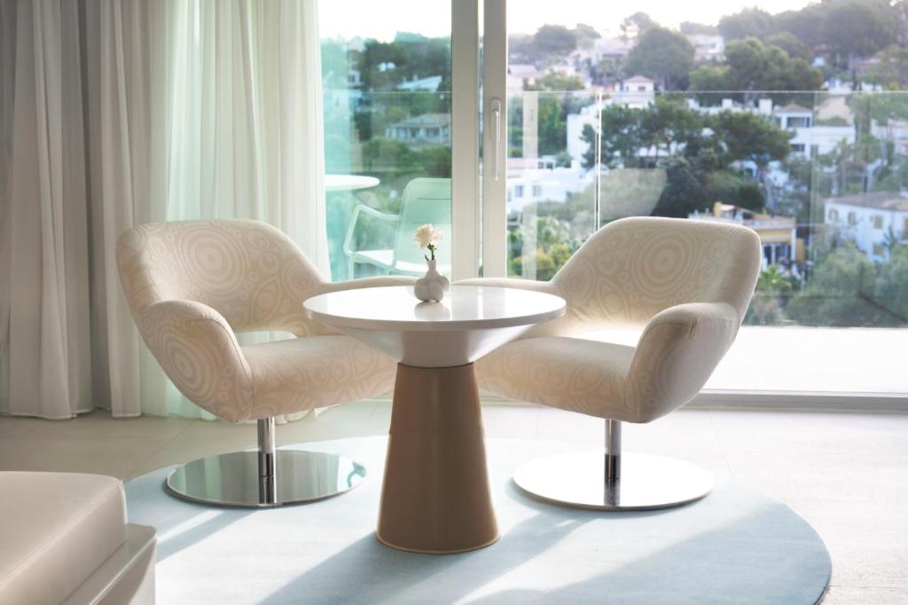 Mūsdienīgs dizaina stūrītis ar divām stilīgām krēsliem un apaļu galdu, kas novietots pie liela loga ar skatu uz dārzu. Gaismas un krāsu harmonija rada mājīgu atmosfēru.