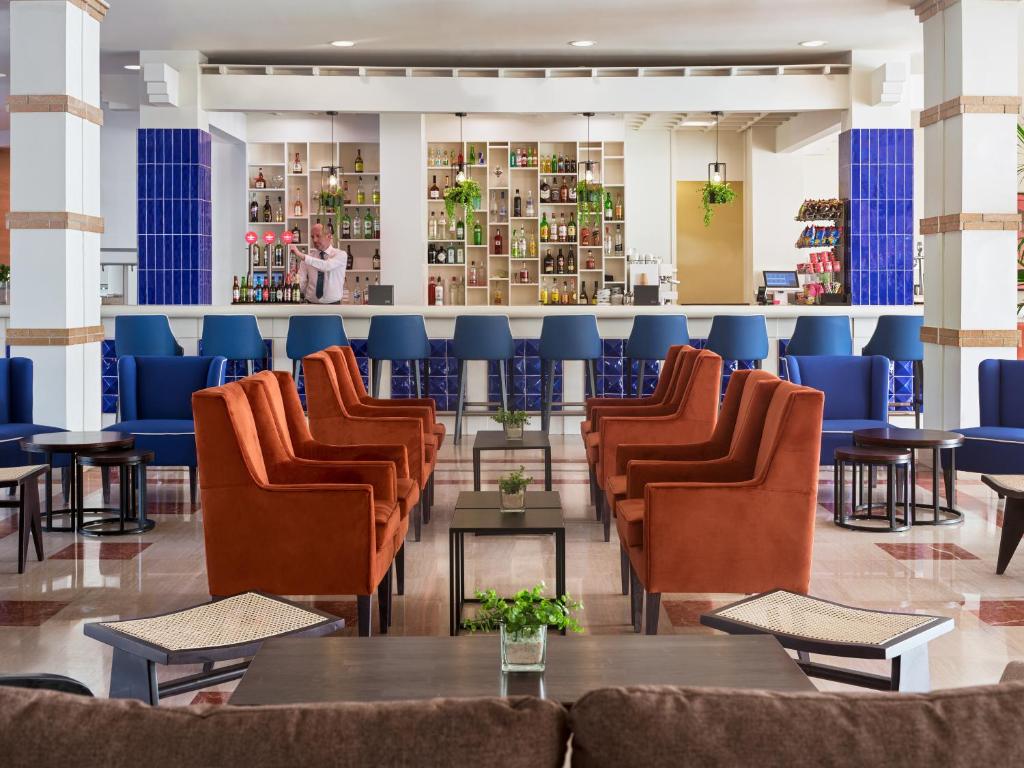 Mūsdienīgs viesnīcas bārs ar stilīgām oranžām un zilām krēslām, elegantu galdu un plašu alkoholisko dzērienu klāstu. Telpa ir gaiša un gaumīga, radot patīkamu atmosfēru atpūtai.