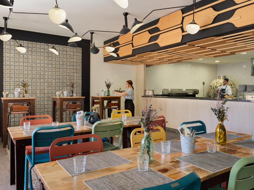 Moderns restorāns ar krāsainām mēbelēm, koka dekoru un stilīgu apgaismojumu, kur viesmīles apkalpo apmeklētājus. Interjerā redzami galdi ar ziediem un eleganti trauki.