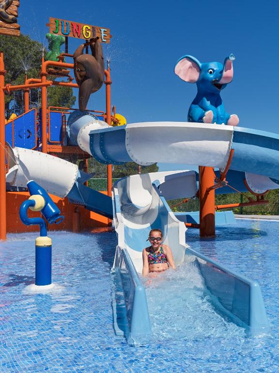 Ūdens slidkalniņš ar jautriem dzīvnieku tēliem un bērnu, kas izbauda braucienu. Spilgti zils debesis un dzidrs ūdens veido priecīgu atmosfēru.