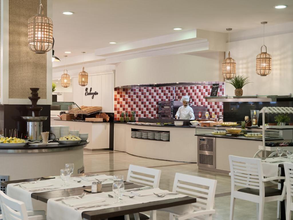 Mūsdienīga restorāna iekārtojums ar atvērtu virtuvi un šefpavāru, kas gatavo ēdienus. Interjerā ir elegants dizains un siltas gaismas, kas rada patīkamu atmosfēru.