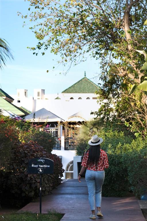 Sieviete pastaigā pa ceļu, kas ved uz Riad Yasmina, ieskauta zaļumiem un ziediem, ar tradicionālu arhitektūru fona. Mierīga atmosfēra un siltie toņi rada relaksējošu noskaņu.