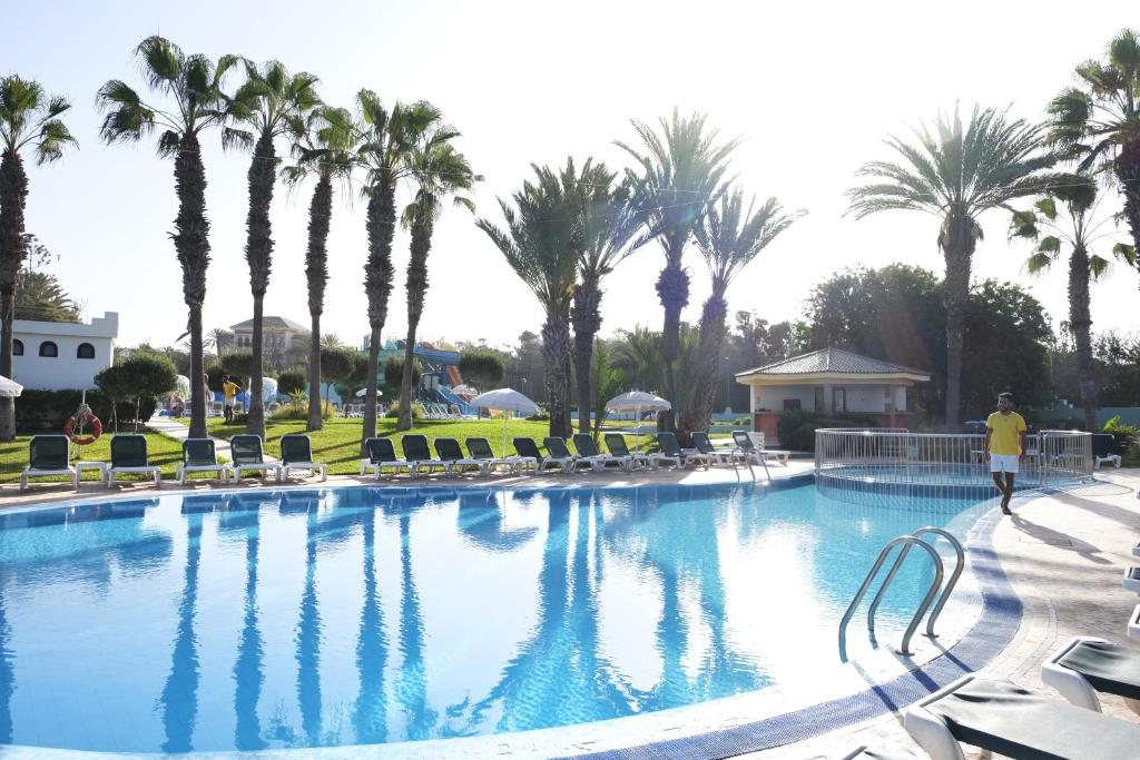 Baseins ar palmām un sauļošanās krēsliem, kas izveidots atpūtas zonā. Aiz baseina redzams ūdens slidkalniņš un zaļa zāle.