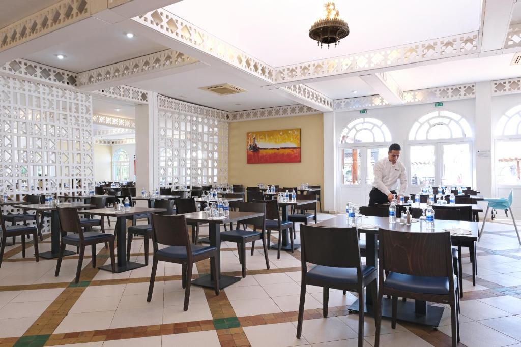 Restorāna interjers ar elegantiem galdiem un krēsliem, kur apkalpojošais personāls sagatavo ēdināšanas telpu. Gaismas un dekorācijas veido patīkamu atmosfēru.