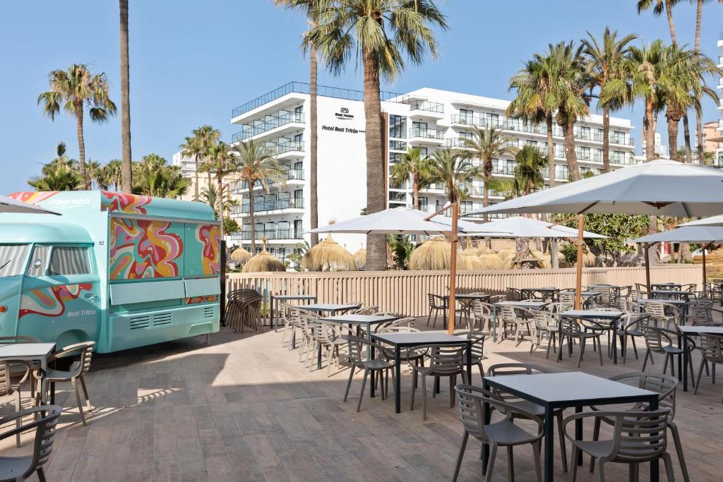 Vasaras terase ar ērtām galdiem un krēsliem, krāsains ēdienu furgons un palmām apkārtnē, skats uz modernu viesnīcu.