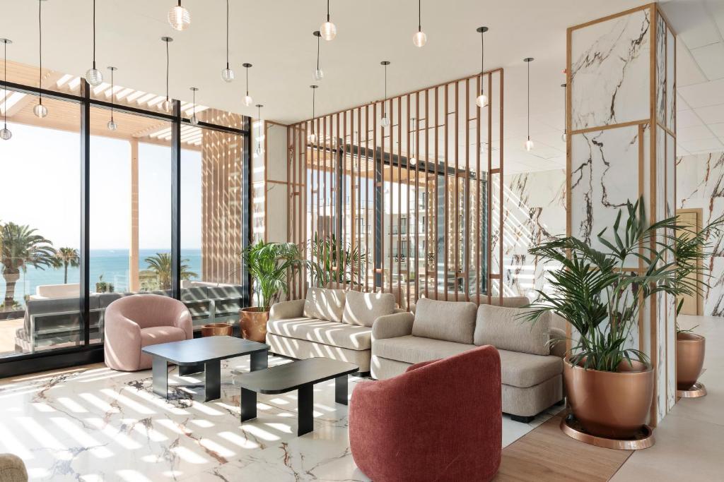 Mūsdienīgs viesnīcas vestibils ar plašiem logiem, kas piedāvā skatu uz jūru, un elegantiem mēbeļu risinājumiem, kas ietver dīvānus un krēslus. Interjers ir apgaismots ar stilīgām laternām un rotāts ar zaļām augiem.