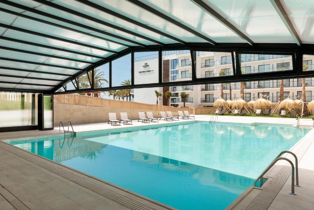 Iekštelpu peldbaseins ar skaidru jumtu un modernu dizainu, apkārt peldbaseinam ir gultas atpūtai. Redzami palmu koki un viesnīcas ēka fonā.