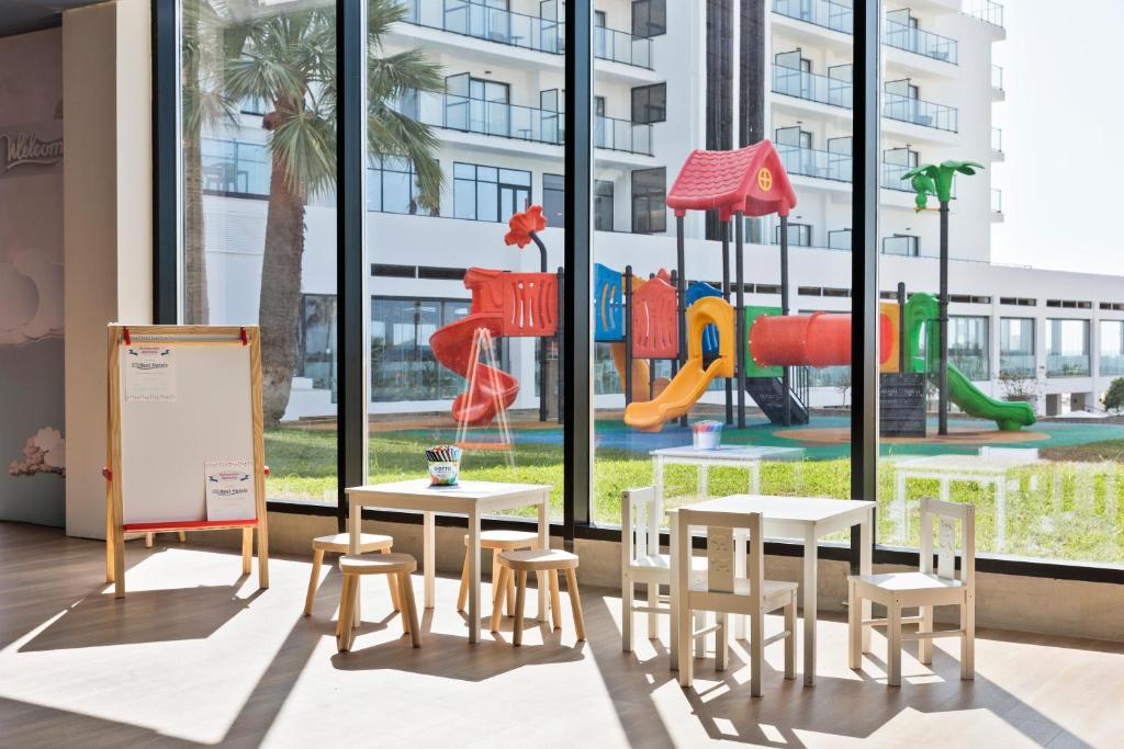 Bērnu rotaļu telpa ar rakstīšanas galdu un krēsliem, caur lielajiem logiem redzams krāsains ārējais rotaļu laukums.