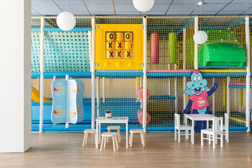 Bērnu rotaļu telpa ar krāsainiem spēļu elementiem, tostarp slidkalniņiem un sietu virsmas. Telpā ir mazi galdi un krēsli, kā arī jautrs karikatūras tēls.