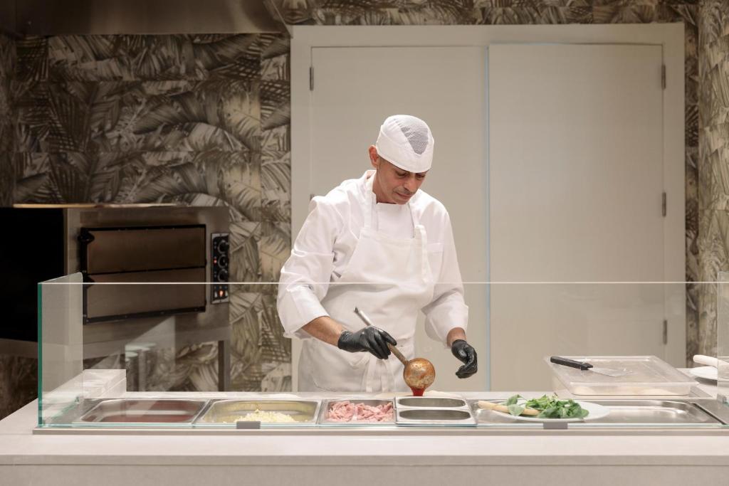 Kāds šefpavārs gatavo ēdienus modernā virtuvē, izmantojot svaigus ingredientus un uzmanīgi strādājot pie stacijas ar stikla ekrānu.