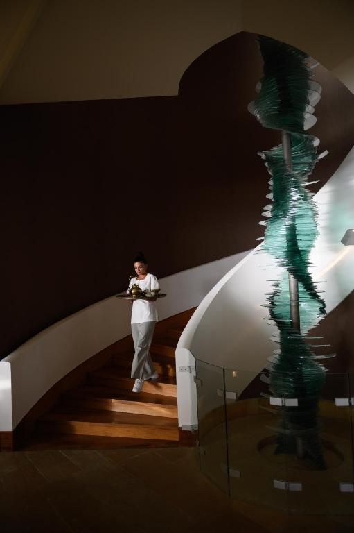 Sieviete kāpj pa spirālveida kāpnēm, nēsājot pasniegšanas paplāti, blakus redzams moderns mākslas objekts no stikla.