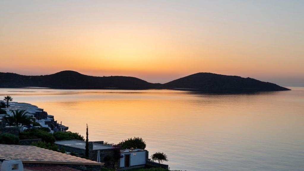 Saulriets virs mierīgas jūras, ar kalniem fona un zaļām ainavām, radot relaksējošu atmosfēru. Ideāla ainava atpūtai un dabas baudīšanai.