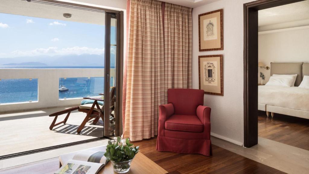Mūsdienīgs viesnīcas numurs ar skatu uz jūru, iekārtots ar sarkanu krēslu un elegantiem mākslas darbiem uz sienas. Atvērti logi nodrošina gaismas plūsmu un piekļuvi balkona zonai.