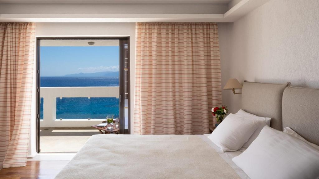 Mūsdienīga viesnīcas istaba ar skatu uz jūru un balkonu, kas ietver gultu ar baltām gultasveļām un mīkstām spilveniem. Aiz loga redzama saulaina piekraste un zila jūra.