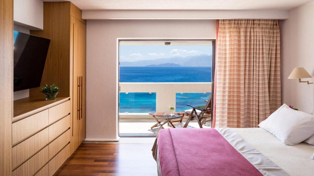 Mūsdienīgs guļamistabas interjers ar skatu uz jūru, kurā ir ērta gulta, televizors un koka mēbeles. Logā redzamas terases mēbeles, kas aicina baudīt brīvdienu atmosfēru.