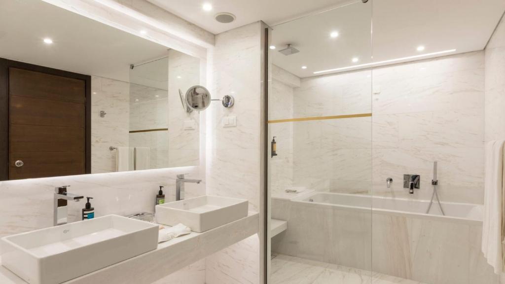 Mūsdienīgs vannas istabas interjers ar gaišām marmora flīzēm, divām izlietnēm un stikla dušu. Telpā ir daudz gaismas un eleganta dizaina elementi, kas nodrošina komfortu un stilīgumu.