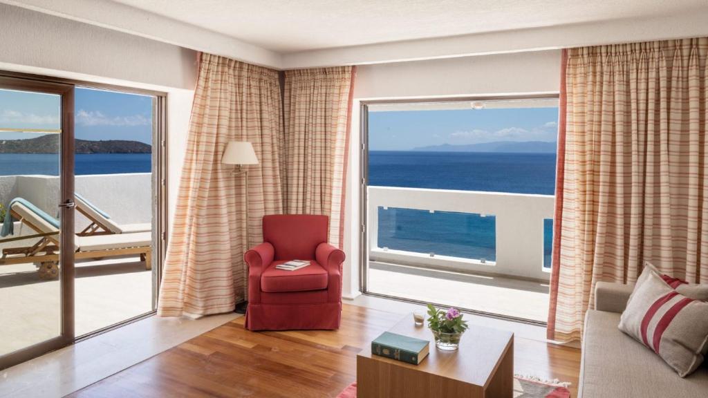 Gaiša viesnīcas istaba ar skatu uz jūru, kurā redzams sarkans krēsls, koka galds ar ziediem un dīvāns, kā arī lielas logu durvis ar aizkariem.