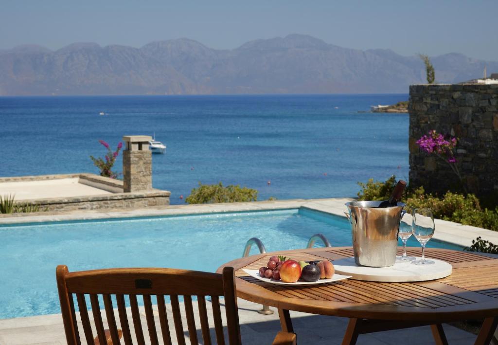 Peldbaseins ar skatu uz jūru, uz galda ir augļi un šampanieša pudele. Apkārt ir skaisti kalni un ziedi.