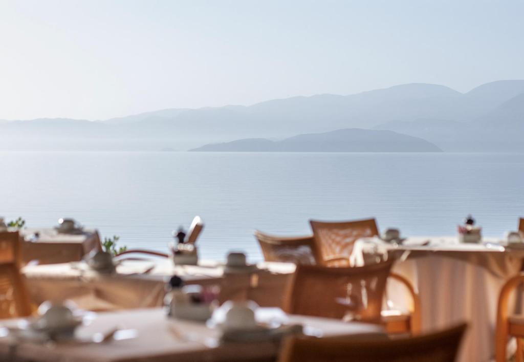 Mierīgas ainavas skats no restorāna ar galdiem un ūdens virsmu fonā, kas atspoguļo kalnus.