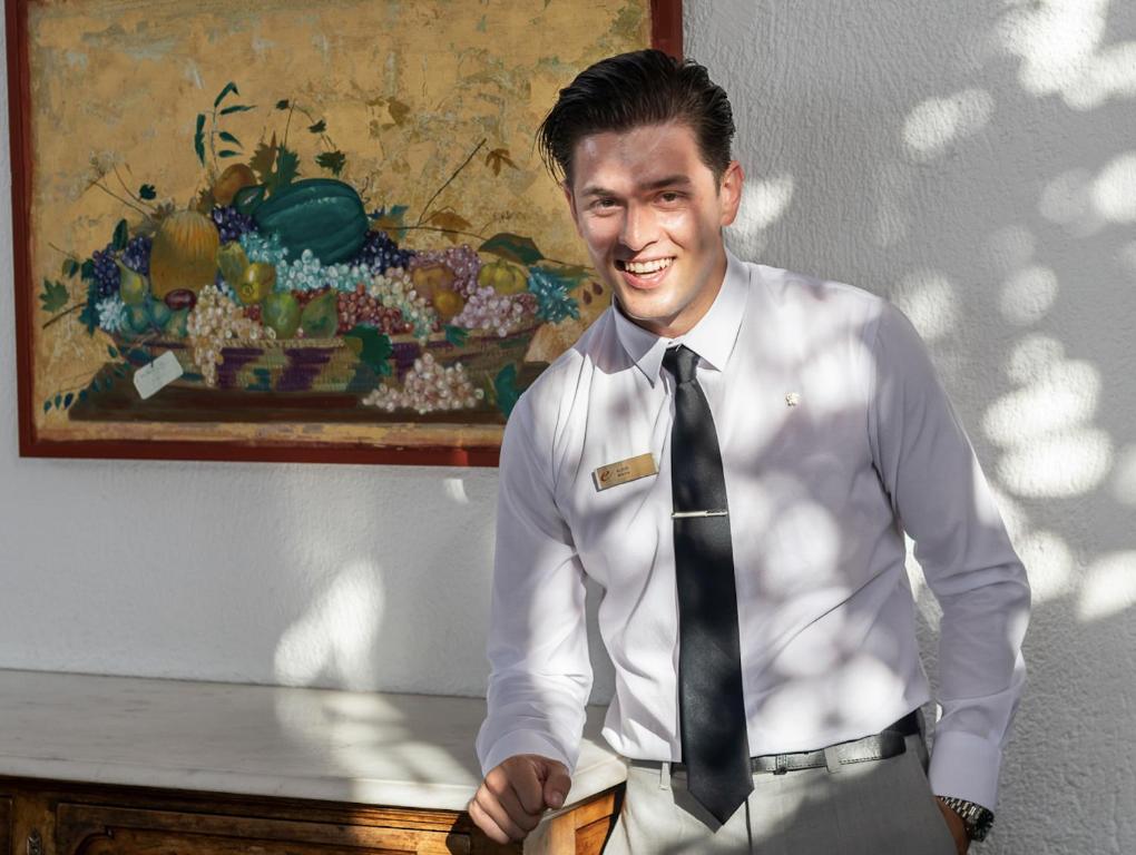 Jauns viesmīlis stāv pie galda, smaidošs un eleganti ģērbts, ar šķērsām saules gaismām, kas krīt uz viņu un uz krāsainu augļu gleznu aiz viņa.